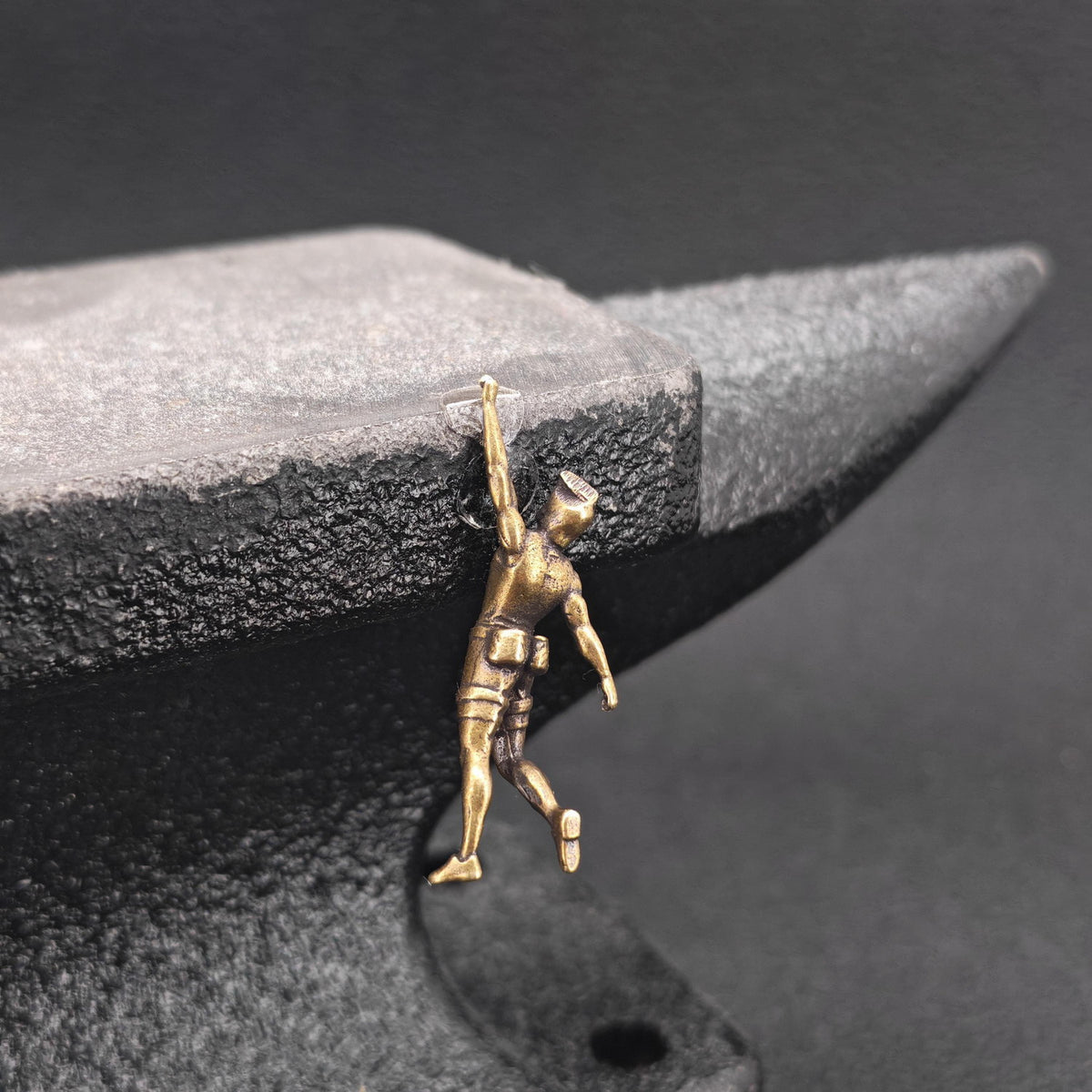 A small gold-toned metal pendant shaped like a worker climbing or leaning against a black anvil, photographed close-up against a dark textured background.