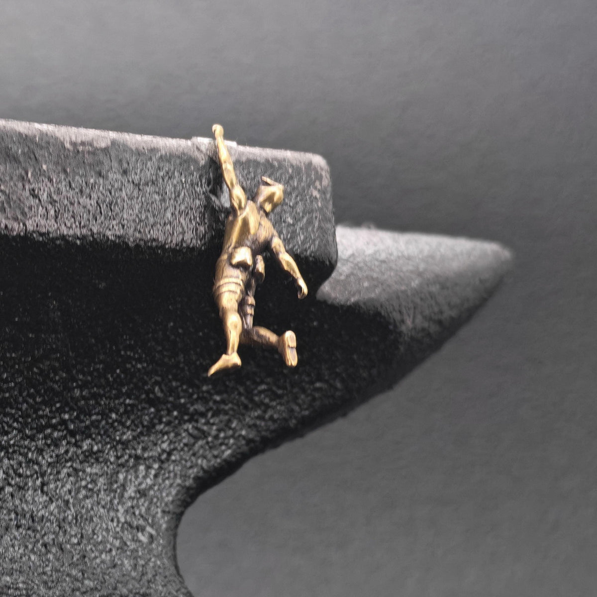A small gold-toned pendant or charm shaped like a person climbing or hanging, displayed on a dark textured jewelry stand against a dark gray background.