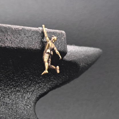 A small gold-toned pendant or charm shaped like a person climbing or hanging, displayed on a dark textured jewelry stand against a dark gray background.
