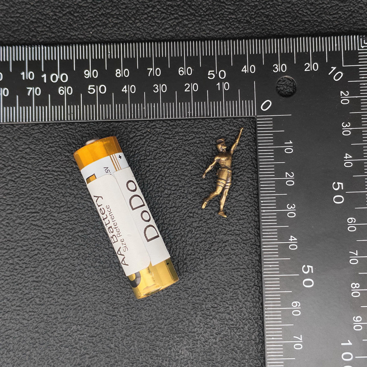 A small metallic gold figurine of a child standing on a black textured surface, shown next to a yellow and white Dodo brand cylindrical item and metal rulers for scale.