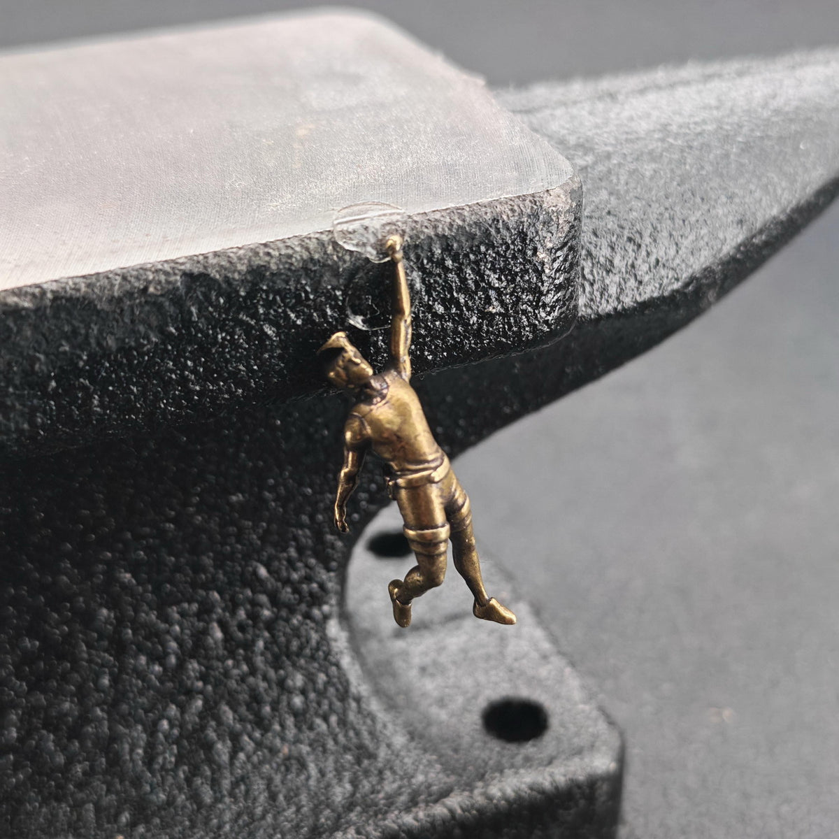 A small bronze-finished figurine of a gymnast hanging from a black anvil, photographed in close-up against a dark gray background.